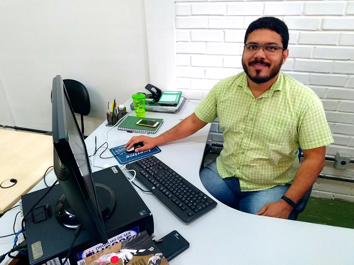 Walber Santana, da coordenadoria de sustentabilidade da UFRPE, sentado em frente ao computador. Camisa verde, calças jeans